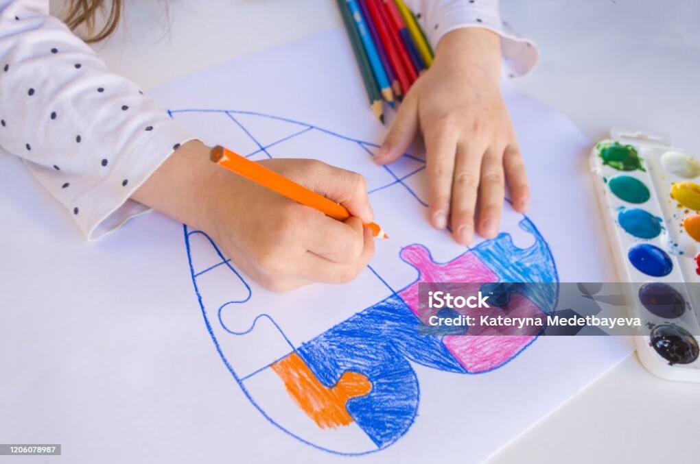 A child is drawing colorful heart with jigsaw puzzle on white background as a symbol of autism, with colorful pencils and watercolors.