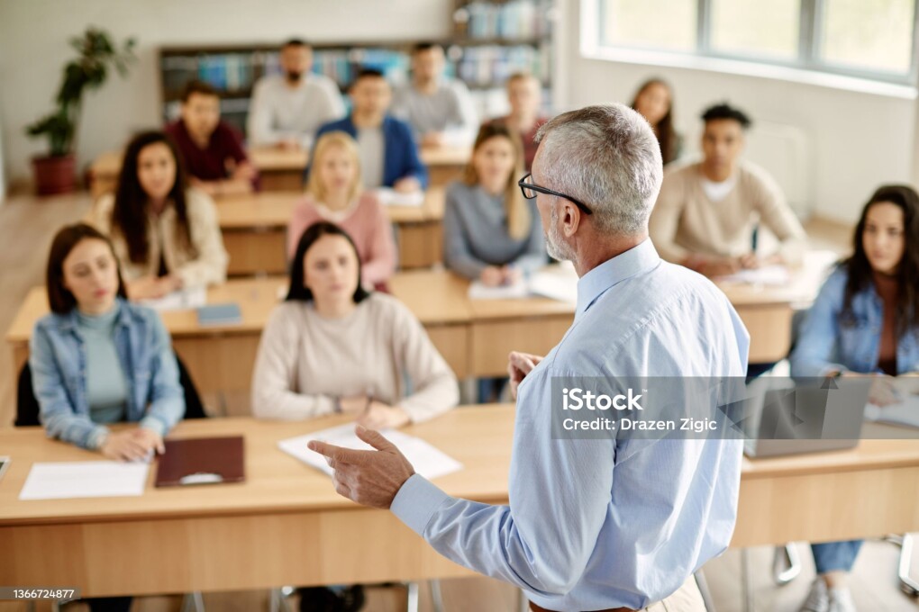 Back view of mature professor giving lecture to large group of college students in the classroom.