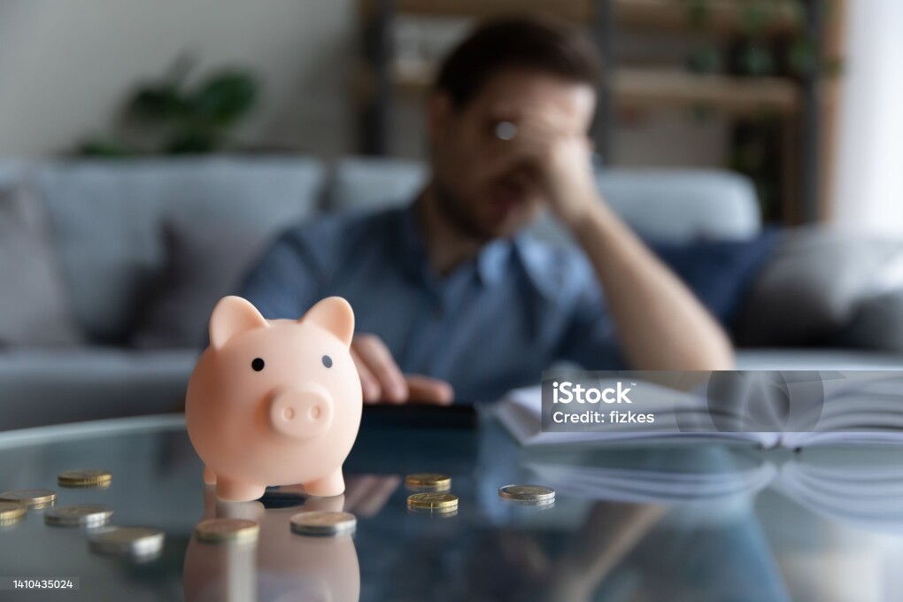 Close up focus on piggybank on table, young man feeling stressed calculating expenditures or taxes, managing future payments or planning investments, suffering from lack of money, accounting concept.