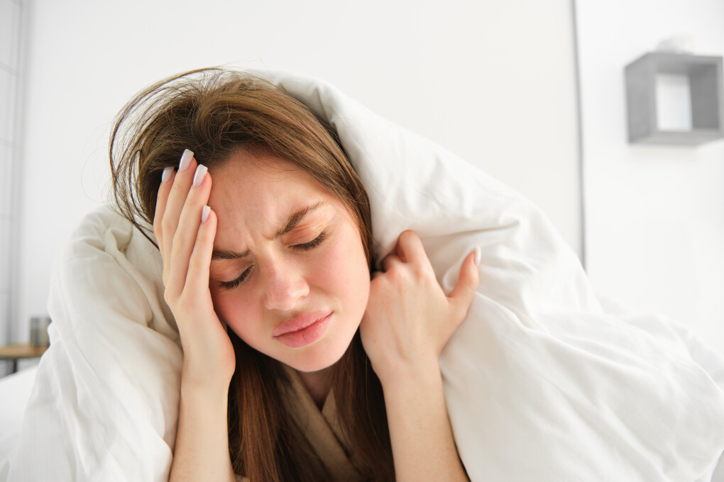 Close up of woman feeling unwell at home, lying in bed under white blankets and frowning, touching head, has headache, migraine, recovering from covid.