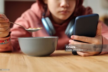 Hand of teenage girl with mobile phone having cornflakes with milk from bowl while sitting by table in front of camera and reading messages