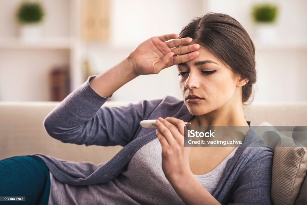 Young woman is looking at the thermometer. She has fever.