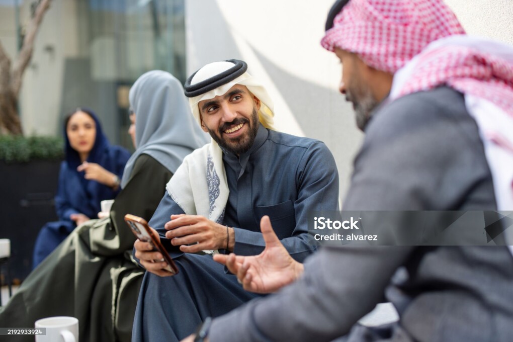 Middle Eastern businessmen in traditional dress talking during a coffee break and looking at a smartphone. Saudi professionals engaging in a discussion at the office cafeteria over a mobile phone.