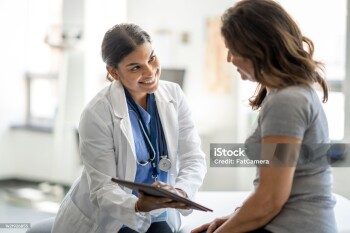 A female doctor of Middle Eastern decent holds out a tablet as she takes notes during a medical appointment with a senior patient.  She is wearing blue scrubs and has a white lab coat overtop, and the patient is dressed casually and has her back to the camera.