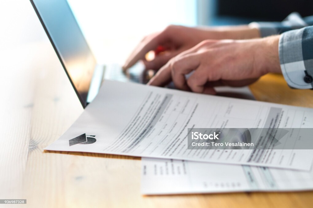 Young man writing college or university application form with laptop. Student applying to school. Scholarship document, admission paper or letter on table. Typing email. Education and communication.