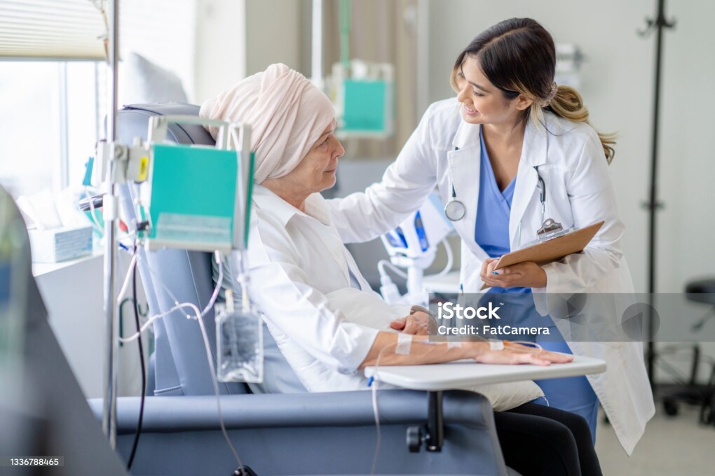 A young female oncologist doctor is checking in on a senior patient of hers who has cancer. She has one hand on her patient's shoulder as she is providing her with moral encouragement during chemotherapy treatment.