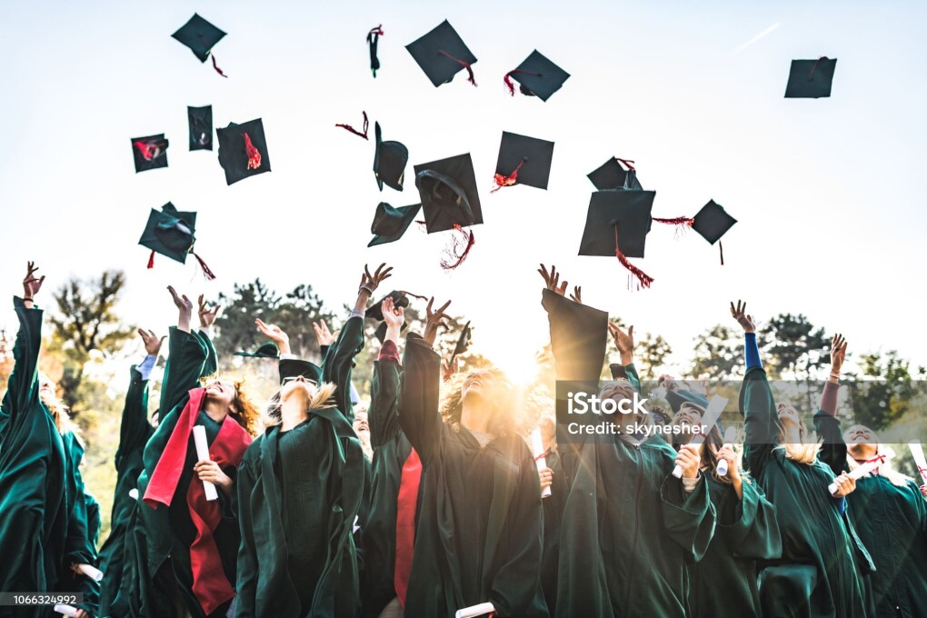 Large group of happy college students celebrating their graduation day outdoors while throwing their caps up in the air.