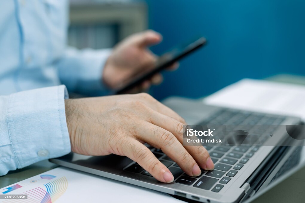 Businessman is typing on a laptop while holding a cell phone. Concept of multitasking and productivity, as the man is able to work on his laptop while also staying connected to his phone