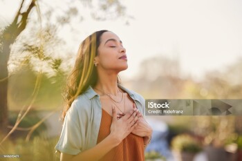 Young latin woman with hand on chest breathing in fresh air in a beautiful garden during sunset. Healthy mexican girl enjoying nature while meditating during morning exercise routine with closed eyes. Mindfulness woman enjoying morning ritual and relaxing technique.