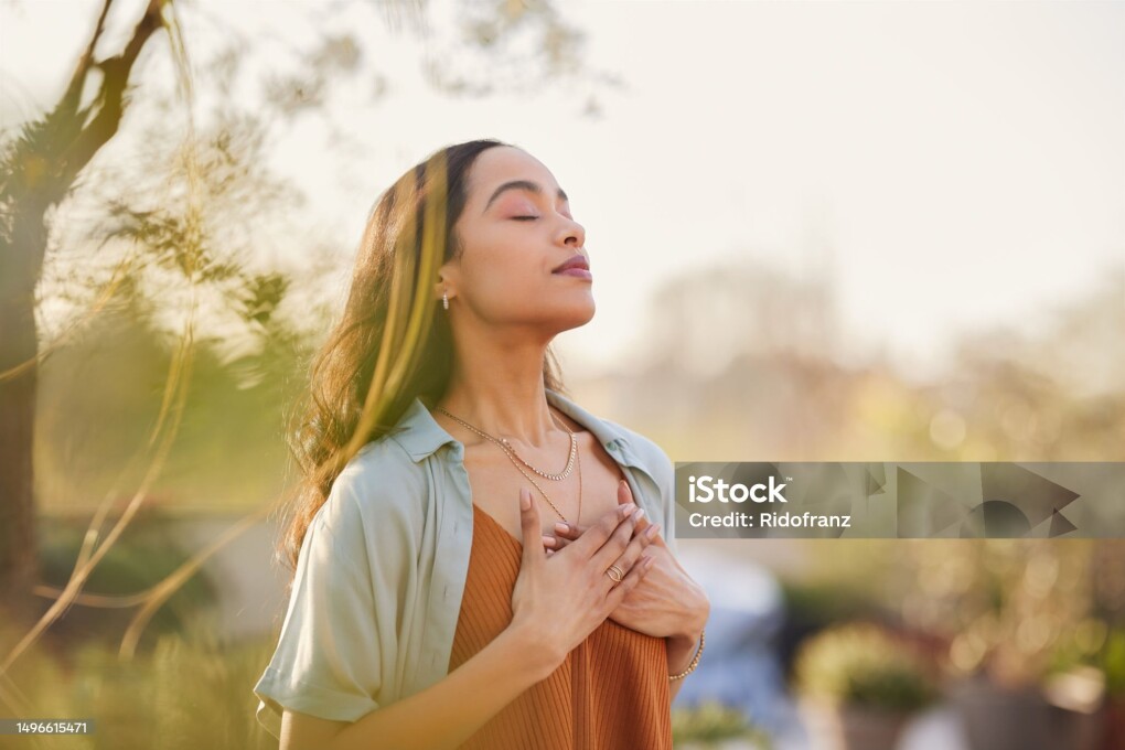 Young latin woman with hand on chest breathing in fresh air in a beautiful garden during sunset. Healthy mexican girl enjoying nature while meditating during morning exercise routine with closed eyes. Mindfulness woman enjoying morning ritual and relaxing technique.