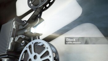 A close up photo of a vintage film projector. The projector is in focus, and the light beam it emits is brightly shining forward. The projector is positioned in front of a light gray background. The image is taken from a low angle, giving the viewer a front-facing perspective of the machine.