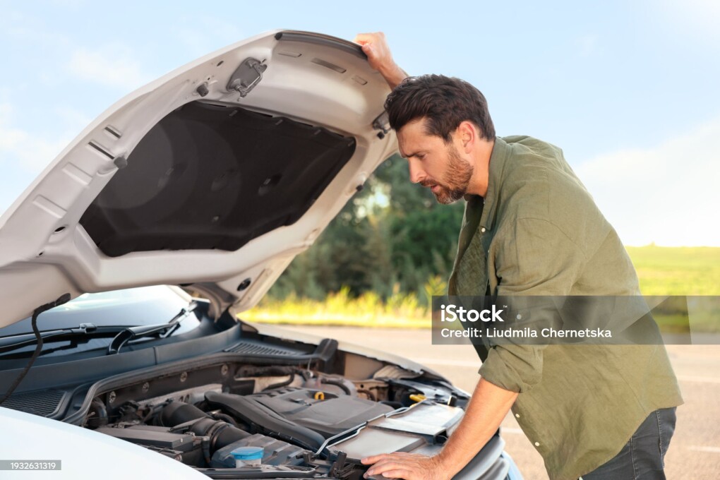 Upset man looking under hood of broken car outdoors