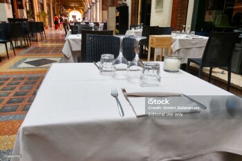 Bologna, Italy, May 16, 2024: Elegant outdoor dining setup in a picturesque european alley, featuring a neatly arranged white tablecloth, glassware, cutlery, and a candle holder on a tiled floor background. ideal for themes related to summer dining, travel, and european vacation.