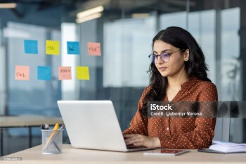 A dedicated office worker is focused on her laptop, surrounded by sticky notes on a glass wall. Her concentration highlights productivity and organization in a modern office setting.