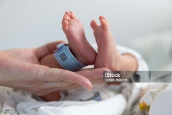 A baby's feet are held up by a person. The baby is wearing a blue tag. The baby is in a hospital bed