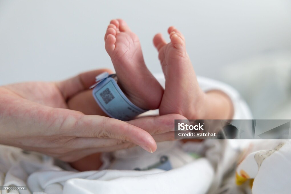 A baby's feet are held up by a person. The baby is wearing a blue tag. The baby is in a hospital bed