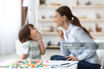 Speech training for kids. Professional woman specialist training with little boy at cabinet, teaching him right articulation exercises, side view