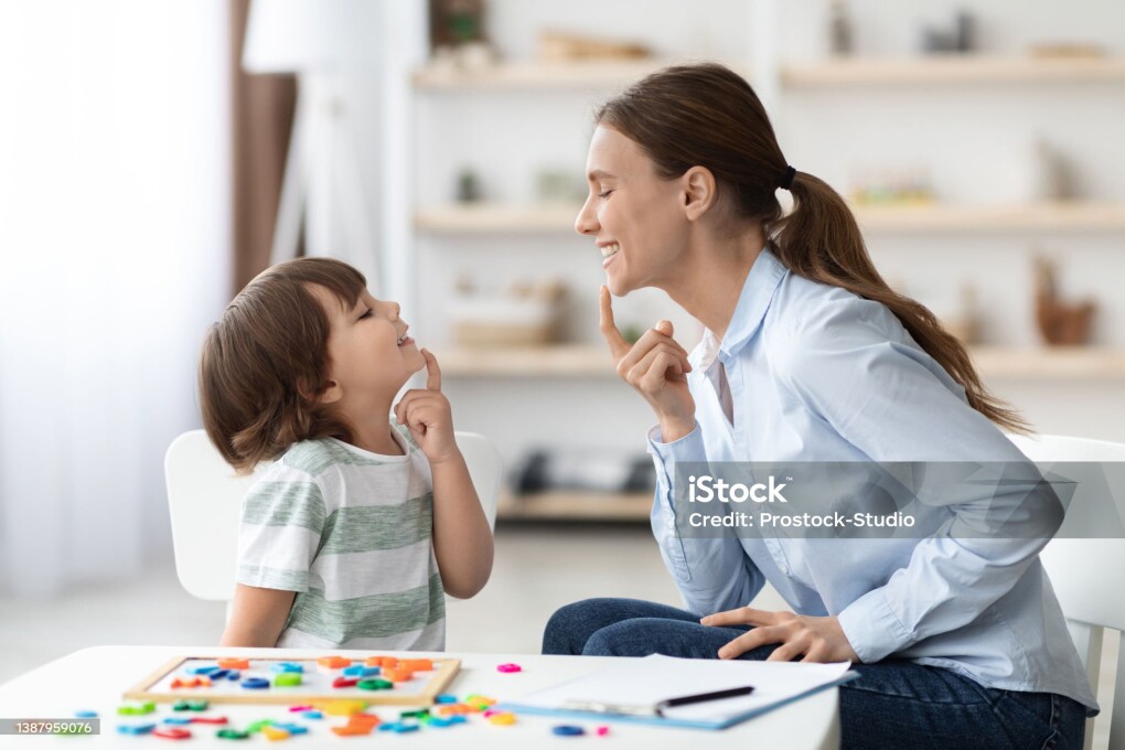 Speech training for kids. Professional woman specialist training with little boy at cabinet, teaching him right articulation exercises, side view