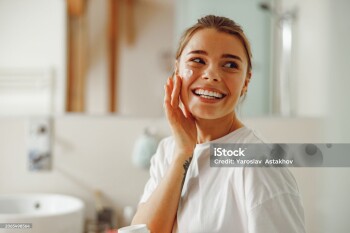 Happy woman applying hydrating moisturizer on her face sitting in bathroom. Home beauty routine