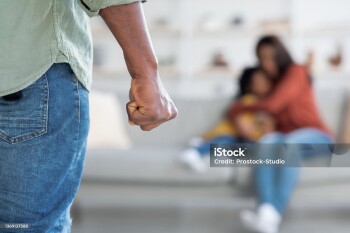 Domestic Violence. Unrecognizable African American Man Threatening Wife And Daughter With His Fist, Scared Mother Embracing Little Girl While Sitting Together On Couch, Selective Focus On Male Hand