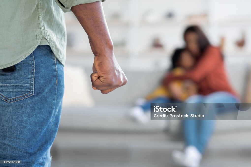 Domestic Violence. Unrecognizable African American Man Threatening Wife And Daughter With His Fist, Scared Mother Embracing Little Girl While Sitting Together On Couch, Selective Focus On Male Hand