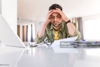 Photo of disappointed young man working on laptop at home and going through paperwork.