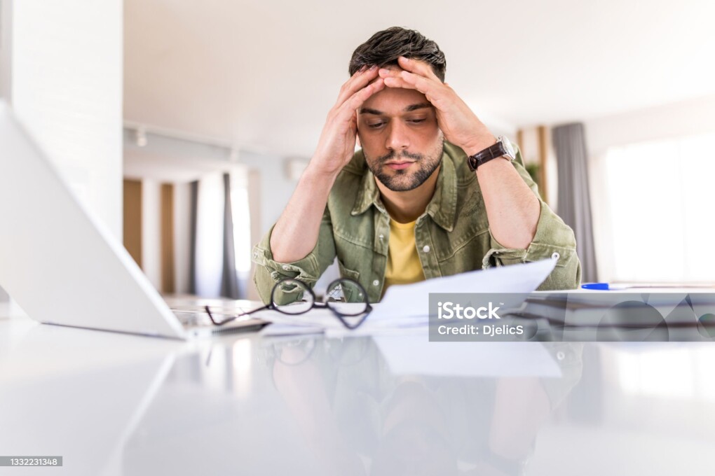 Photo of disappointed young man working on laptop at home and going through paperwork.