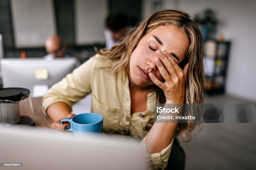 Tired and exhausted young female entrepreneur drinking coffee and rubbing eyes while feeling stressed and worried in coworking space
