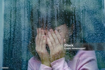 Sad child is covering her face with hands and crying behind rainy window