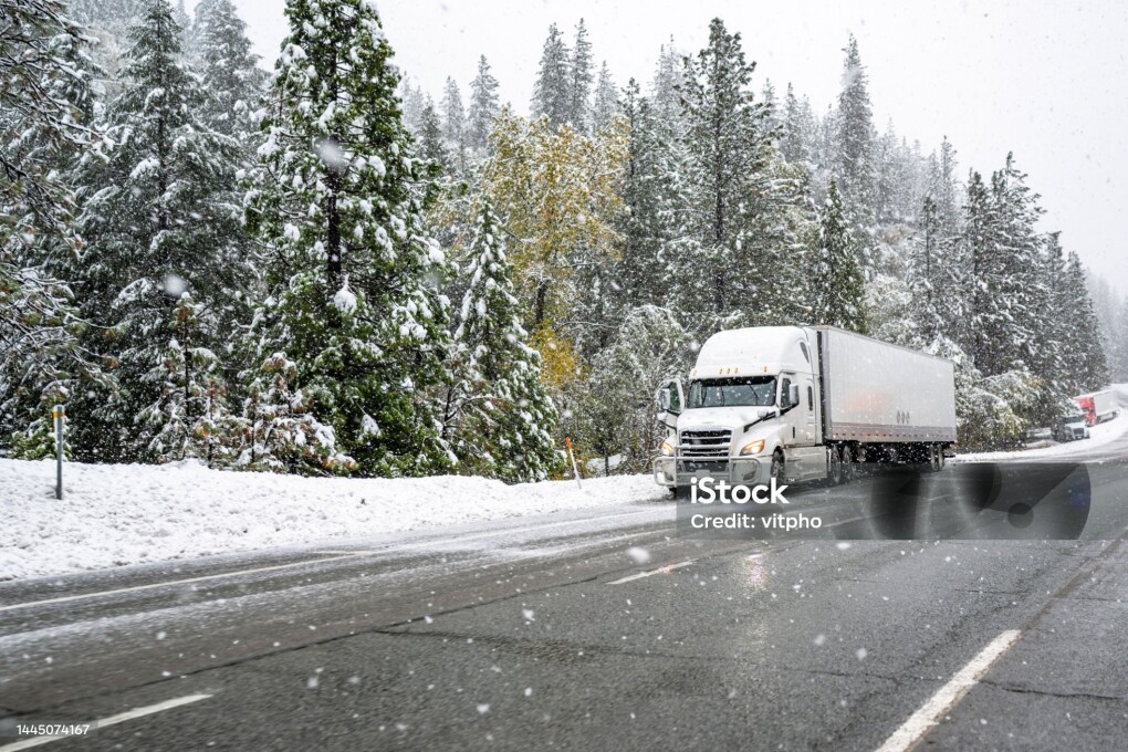 White big rig industrial semi truck with grille guard transporting cargo in dry van semi trailer standing on road shoulder of a winter highway during a snow storm near Shasta Lake in California