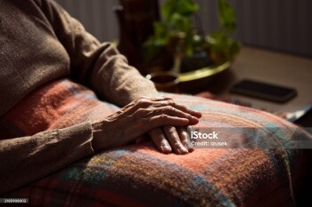 Senior woman sitting in armchair at home with legs covered by blanket. Close up of hands, unrecognizable person.