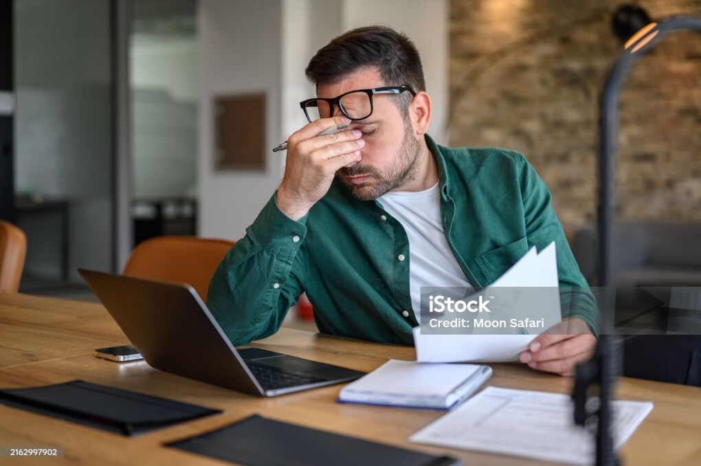 Anxious male financial advisor rubbing eyes while working over laptop and analyzing reports at desk