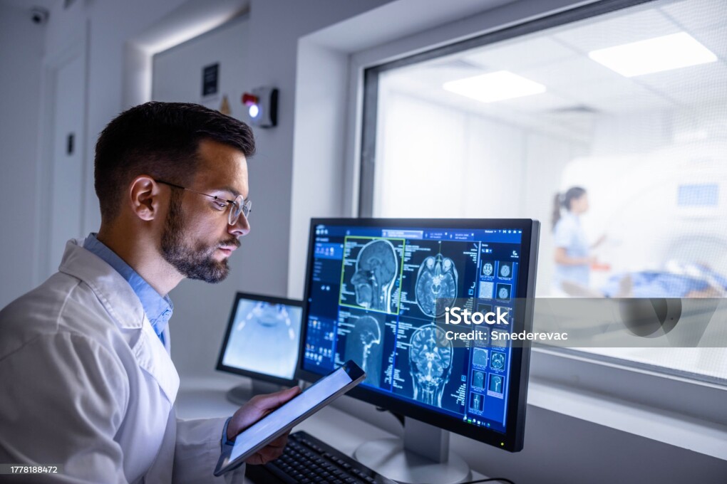 Doctor examining X-ray images on display in control room while in background nurse preparing the patient for new MRI scanning test.
