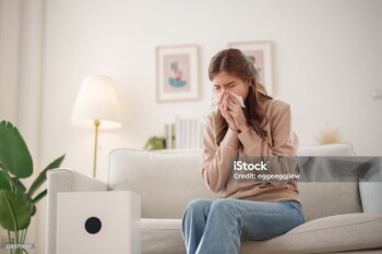 Young Asian woman sits on a couch, sneezing and holding a tissue to her nose while an air purifier operates nearby to improve air quality.