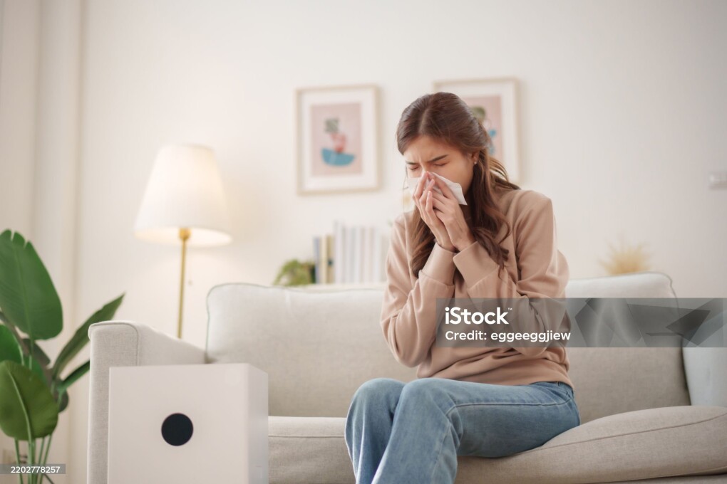 Young Asian woman sits on a couch, sneezing and holding a tissue to her nose while an air purifier operates nearby to improve air quality.