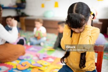 Preschool girl covers her ears and closes her eyes while other kindergarten children play loudly behind her. Concept of noise sensitivity, sensory overload, and emotional response in early childhood