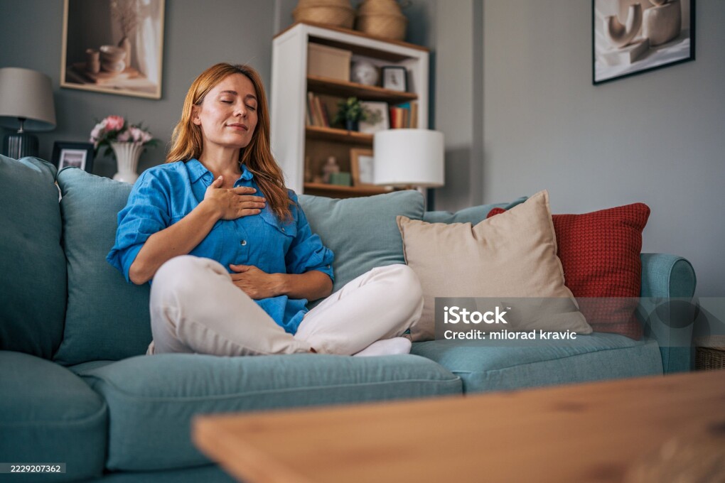 Mid adult woman with closed eyes is sitting in lotus position on a comfortable sofa, practicing mindfulness and enjoying a moment of peace and relaxation in her living room