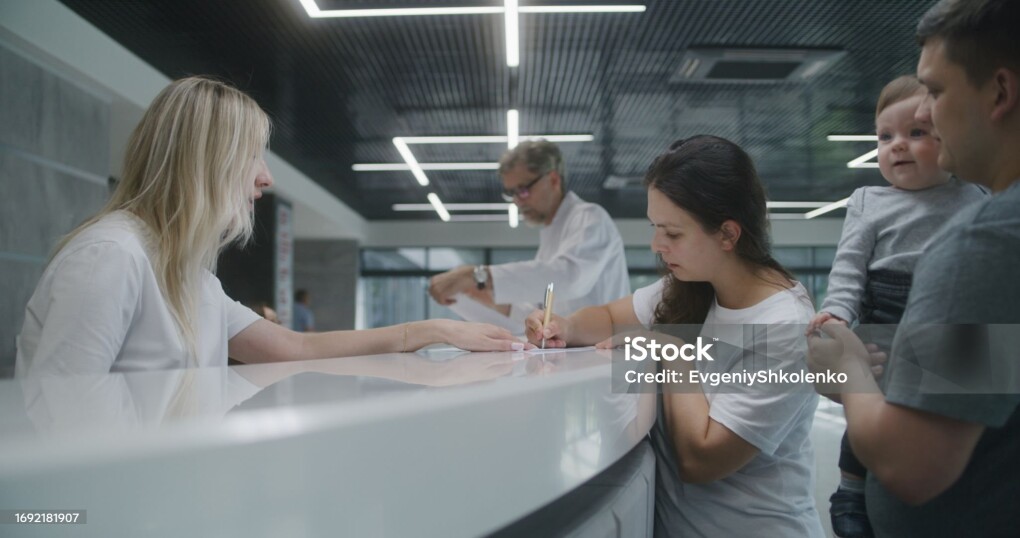 Family with little child stands near reception desk in clinic lobby area. Woman fills out documents and papers, makes appointment with doctor. Medical staff work in modern medical center. Health care.