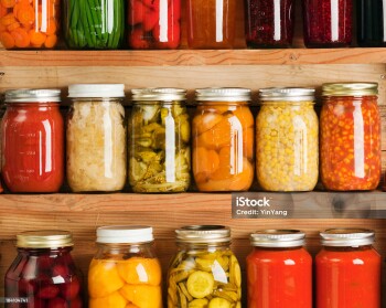Wooden storage shelves holding home canning vegetable and fruit preserves varieties, lined up in rows of glass jars. Canned food staple include jellies, sauces, carrot slices, green beans, tomatoes, corn, sweet potatoes, sauerkraut, roasted red peppers, dill pickles, red beets, peaches, rasperry jam, orange marmalade, grape jelly, tomato paste and corn soup.