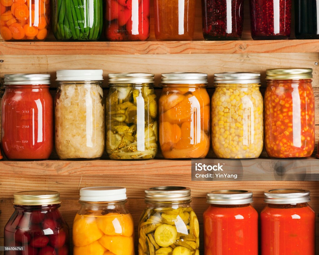 Wooden storage shelves holding home canning vegetable and fruit preserves varieties, lined up in rows of glass jars. Canned food staple include jellies, sauces, carrot slices, green beans, tomatoes, corn, sweet potatoes, sauerkraut, roasted red peppers, dill pickles, red beets, peaches, rasperry jam, orange marmalade, grape jelly, tomato paste and corn soup.