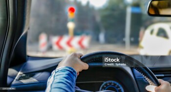 Hands holding a car's black steering wheel. The person is wearing a blue sleeve. The vehicle is stopped at a traffic light, visible through the windshield with a red signal illuminated. In the background, traffic signs with chevron patterns and other blurred vehicles are visible. The dashboard has a visible air vent and part of the instrument panel. The interior suggests a modern car design.