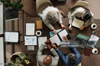 Group of middle aged multiethnic business professionals collaborating around table, reviewing documents and using laptop, top view showing teamwork and corporate meeting environment