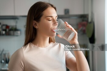 Thirsty young woman drinking fresh water from glass. Home office kitchen interior. Headshot portrait. Dehydration prevention, normal bowel function and balance of body maintenance concept