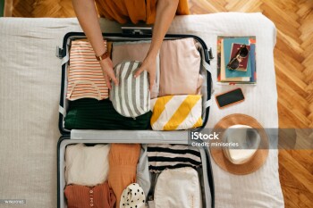Shot from above of an anonymous woman packing things in her suitcase on the bed. She is holding and putting a grey and white stripped cosmetics bag in. There is her mobile phone, books and sunglasses on the bed.