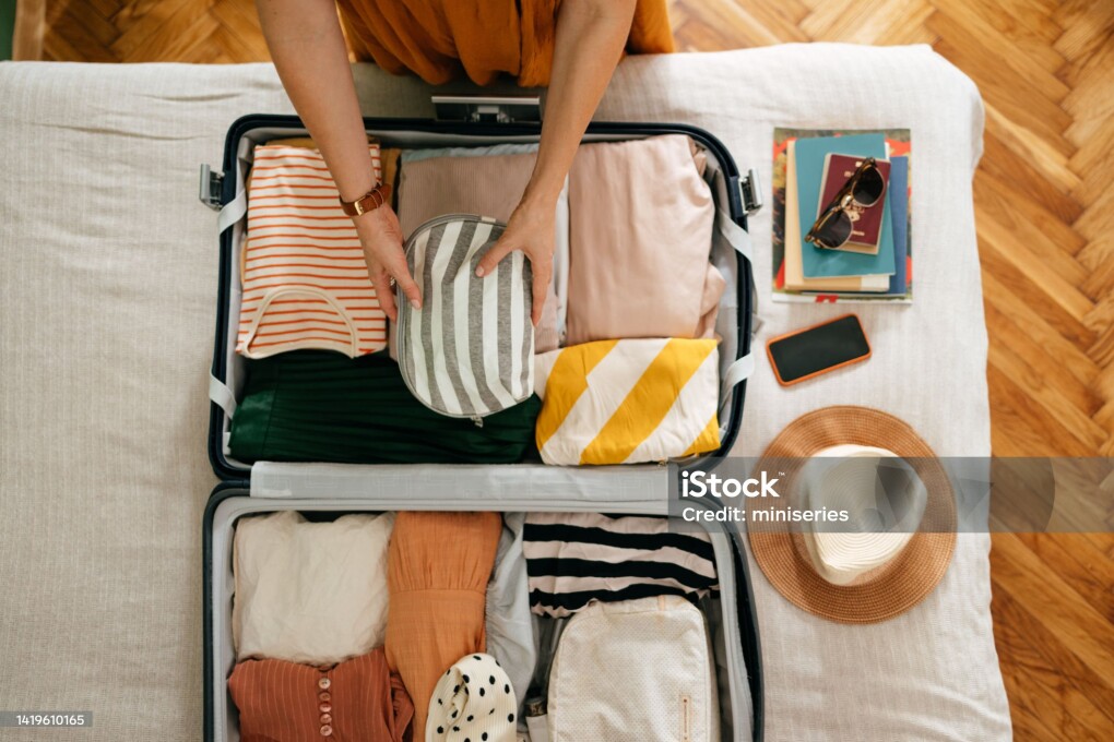 Shot from above of an anonymous woman packing things in her suitcase on the bed. She is holding and putting a grey and white stripped cosmetics bag in. There is her mobile phone, books and sunglasses on the bed.