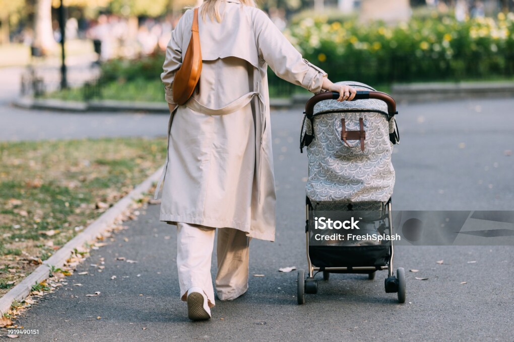 Follow the path of motherhood through the park as a casually dressed mom walks with her baby in the stroller, the rear view revealing the beauty of the shared journey.