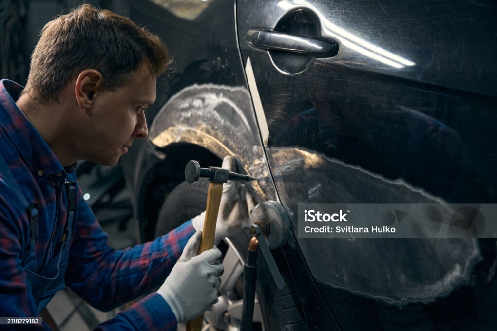 Professional male in protective costume standing near automobile, holding hammer and remove paint, processes metal