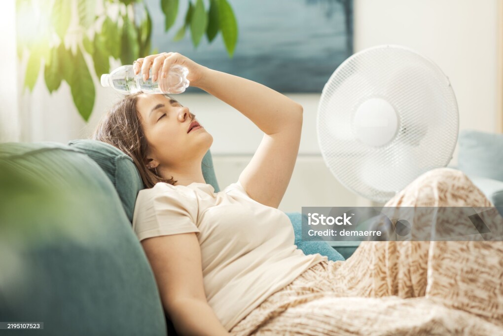 Young woman sitting on the couch at home and keeping a fresh water bottle on her forehead, she is suffering from the heat