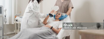 Asian woman on a treatment bed at aesthetic clinic, examining her face in a handheld mirror after receiving a beauty injection. Skincare and anti-aging treatments.