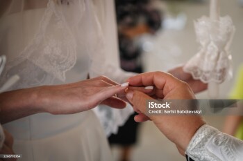 Close-up capturing a sacred wedding moment in an Orthodox church. A priest's hand gently places a gold wedding ring on the bride's finger during the religious sacrament.